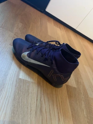 Lila Nike Mercurial fotbollsskor - Säljer ett par lila Nike Mercurial fotbollsskor med högt skaft och silverfärgad swoosh på sidan. Skorna har snörning och dobbar i plast för bra grepp på planen. Materialet är syntet och skon har en cool design med 'KM' tryckt på hälen som står för Kylian Mbappe. Skorna går att få renare men har inte haft ork för det. 