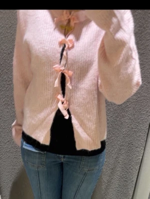 Ljusrosa kofta med rosetter - Supersöt ljusrosa kofta i stickat material med fyra dekorativa rosetter framtill. Koftan har en loose passform och långa ärmar, perfekt för dig som gillar en mysig och trendig look. Passar lika bra till jeans som över en klänning.