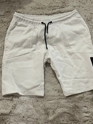 Vita Peak Performance shorts med snörning - Säljer ett par vita shorts från Peak Performance med svart snörning i midjan och en svart patch med logga på ena benet. Shortsen har fickor fram och en bakficka, tillverkade i mjuk bomull för en chill och sportig vibe.