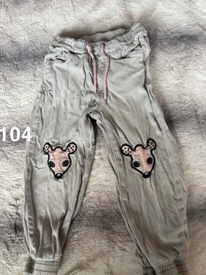 Grå byxor med djurmotiv - Grå byxor med resår i midjan och rosa snörning. Byxorna har söta broderade djurmotiv på knäna och muddar vid bensluten. Perfekta för chill dagar eller lek. 🌸 Fint använt skick. Storlek 104