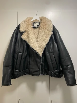 Svart ”skinn”jacka från H&M med fuskpäls - Säljer en svart teddyjacka från H&M med stor krage och foder i beige fuskpäls. Jackan har dragkedja, bälte nertill och detaljer vid ärmslut. Oversized passform och tillverkad i polyester och syntetmaterial. Perfekt för kalla dagar när du vill vara både varm och snygg.