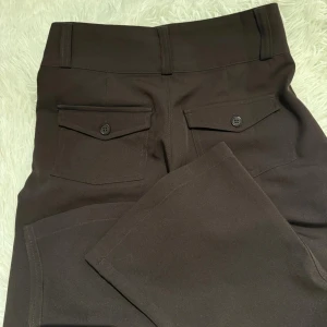 Bruna kostymbyxor med ficklock - Mörkbruna kostymbyxor med raka ben och två bakfickor med ficklock och knappar. Byxorna har hög midja, bälteshällor och stängs med två knappar framtill. Snygg klassisk look som passar till skjorta eller stickad tröja.