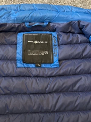 Blå pufferjacka från Sail Racing - Säljer en blå pufferjacka från Sail Racing med coolt badge på ärmen och två dragkedjeförsedda fickor framtill. Jackan har ståkrage och quiltad design som ger en sportig vibe. Perfekt för dig som vill ha en lätt och smidig jacka till kyligare dagar.pris går att diskutera vid snabb affär.