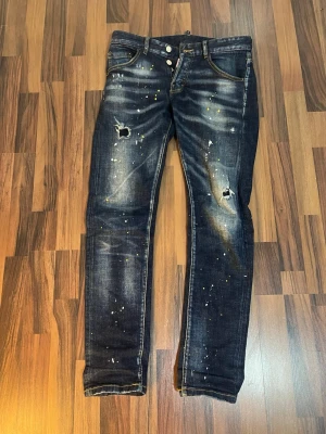 Dsquared2 blå jeans - Säljer ett par blå jeans från Dsquared2 med coola slitningar och färgstänk i vitt och gult. Jeansen har klassisk femficksmodell, gul kontrastsöm och patch bak i midjan. Rå och trendig look med knappgylf. Knappt använda. Skulle säga att dom är 32/32 i storlek. Är 172 å dom passar perfekt, skulle säga att dom passar på dom som e 165-177cm ungefär