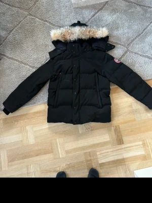 Canada Goose  - Säljer en svart dunjacka från Canada Goose med pälskantad huva och klassisk logga på ärmen. Jackan har långa ärmar, stora fickor framtill och stängs med både dragkedja och knappar. Perfekt för kalla dagar och riktigt snygg vinterstil.