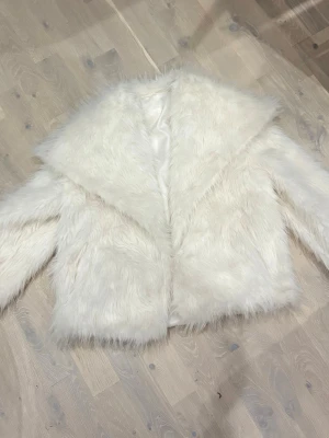 Vit pälsjacka i fuskpäls, stl S - Fluffig vit pälsjacka i fuskpäls med bred krage och lång ärm. Jackan är helvit och har ett mjukt, glansigt innerfoder. Tillverkad i 100% polyester och har en oversized känsla. Perfekt för dig som vill sticka ut och hålla dig varm under kyliga dagar.