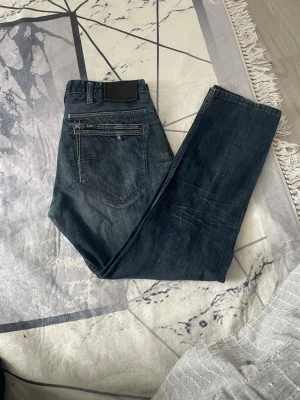 Lee Clark Tapered jeans W31 L32 - Säljer ett par mörkblå Lee Clark Tapered jeans med klassisk femficksdesign och snygga slitningar. Jeansen har en tapered passform som smalnar av nedtill och är tillverkade i slitstarkt denimtyg. Perfekta för dig som gillar en clean och tidlös look.