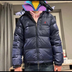 Mörkblå dunjacka från Polo Ralph Lauren - Säljer en mörkblå dunjacka från Polo Ralph Lauren med huva och dragkedja. Jackan har en klassisk puffer-look, lång ärm och ikonisk röd logga på bröstet. Perfekt för kalla dagar och riktigt snygg till jeans eller hoodie.