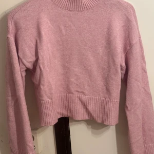 Ljusrosa croppad stickad tröja - Superfin ljusrosa stickad tröja med croppad passform och ribbade detaljer vid hals, ärmslut och nederkant. Perfekt för dig som gillar en enkel men trendig look. Långärmad och mjuk, passar till både jeans och kjol.