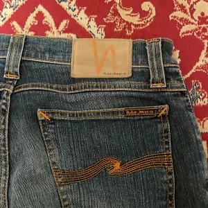 Nudie Jeans - Säljer ett par klassiska blå skinny jeans från Nudie Jeans med snygga slitningar och kontrastsömmar i orange. Jeansen har fem fickor, normal midja och är tillverkade i stretchig denim för en bekväm passform. Perfekta för dig som gillar en smal siluett.