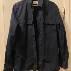 Mörkblå overshirt från Samsøe Samsøe - Snygg mörkblå overshirt från Samsøe Samsøe i bomull. Modellen har två stora bröstfickor med knappar och klassisk krage. 