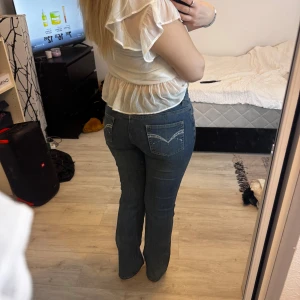  blå bootcut jeans - Säljer ett par klassiska  jeans i blå denim med bootcut passform. Jeansen har låg midja, fem fickor och snygga broderade detaljer på bakfickorna. Perfekta för dig som gillar en tidlös och avslappnad stil.