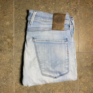 Replay Anbass ljusblå jeans - Säljer ett par Replay Anbass jeans i ljusblå tvätt med klassisk femficksdesign och snygga slitningar på ena benet. Jeansen har tajt passform och raka ben, med Replay-logga på bakfickan och läderpatch i midjan. Perfekta för en avslappnad och trendig look. Priset är  inte hugget i sten!