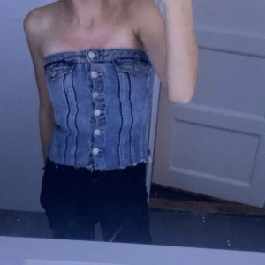 Blå jeans tubtopp med knappar - Säljer en snygg blå tubtopp i jeans med markerade sömmar och stora vita knappar framtill. Toppen har rå kant nertill för en cool look och är perfekt för dig som gillar streetstyle.