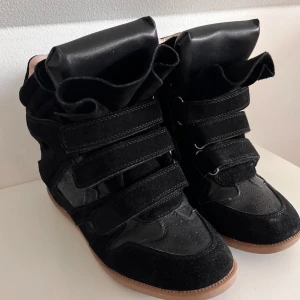  svarta sneakers med kardborre - Svarta sneakers med cool design, högt skaft och breda kardborreband. Skorna är gjorda i mocka och skinn med beige insida och gummisula. Perfekta för dig som gillar statement-sneakers med unik look. 