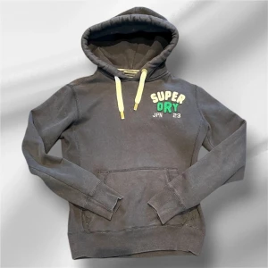 Unik Superdry hoodie - Mörkgrå hoodie från Superdry med broderad logga i vitt och grönt på bröstet. Tröjan har huva med ljusa snören och en klassisk känguruficka framtill. Mjuk insida i fleece och långa ärmar. Perfekt för chill dagar eller när du vill ha en avslappnad stil.