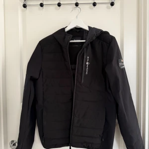 Svart pufferjacka från Sail Racing - Svart pufferjacka från Sail Racing med huva och dragkedja framtill. Jackan har stilren design med logga på bröstet och ärm, quiltade paneler och lång ärm. Perfekt för kyliga dagar och enkel att matcha med allt.