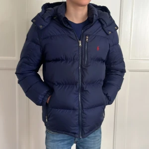 Mörkblå dunjacka från Ralph Lauren - Mörkblå pufferjacka från Ralph Lauren med röd logga på bröstet. Jackan har huva,jackan har en klassisk pufferlook som är både varm och stilren. Perfekt för kalla dagar.ny pris 3295 mitt pris 699 men jackan har några små skavanker, se bilder