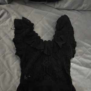 Svart volangblus från Zara - Supersöt svart blus från Zara med virkade detaljer och volanger längs v-ringningen och axlarna. Ärmlös modell med romantisk känsla och stretchig ribbad nederkant. Perfekt att styla med jeans eller kjol för en trendig look.