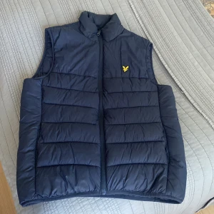 Mörkblå dunväst från Lyle & Scott - Snygg mörkblå dunväst från Lyle & Scott med gul logga på bröstet. Västen har ståkrage, quiltad design och dragkedja framtill. Perfekt för lager-på-lager och enkel att matcha med hoodies eller tröjor.