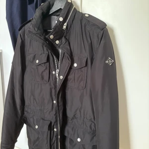 Svart parkajacka från J.Lindeberg - Svart parkajacka från J.Lindeberg med flera fickor framtill, silverfärgade knappar och dragkedja. Jackan har hög krage, axelklaffar och en stilren badge på ärmen. En liten defekt i fickan ett litet hål som ej är märkvärdigt. Perfekt för dig som vill ha en snygg och funktionell jacka med urban känsla.