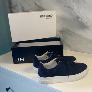 Mörkblå mockasneakers från Selected Homme - Ny pris var 1200 säljer dom för 750, pris kan diskuteras om det är så. Säljer för att dem var för stora och jag har använt dom 1-3 gånger! Snygga mörkblå sneakers från Selected Homme i mjuk mocka med vita, platta sulor och svarta snören. Skorna har en klassisk låg profil och är fodrade med ljus textil på insidan. Perfekta för dig som gillar stilrena och moderna sneakers.
