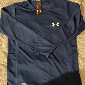 Marinblå långärmad träningströja UA - Snygg marinblå långärmad träningströja från Under Armour med vit logga på bröstet och diskret tryck vid nederkanten. Tillverkad i ett lätt och ventilerande syntetmaterial som passar perfekt till träning. Klassisk rund halsringning och relaxed fit.