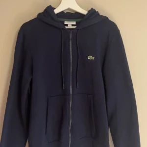 Marinblå hoodie från Lacoste - Säljer en marinblå hoodie från Lacoste i klassisk passform. Tröjan har dragkedja framtill, huva med snörning och en stor ficka på magen. Ikoniska Lacoste-loggan sitter på bröstet. Perfekt för dig som gillar stilren och sportig look. Tröjan är knappt aldrig använd då den har blivit för liten. Nypris: 1795:- (från zalando) 