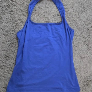 Blå halterneck linnetopp, M - Snygg blå halterneck linnetopp med enkel och stilren design. Toppen har en rundad halsringning och är ärmlös, perfekt för varma dagar. Materialet känns mjukt och lätt, troligen bomull eller polyester. Passformen är normal och toppen är enfärgad utan detaljer.