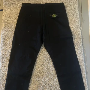 Svarta byxor från Stone Island - Svarta byxor från Stone Island med klassisk patch på bakfickan. Byxorna har snörning i benslut, dragkedjeficka framtill och raka ben. Snygg och stilren design som passar perfekt till streetwear. Materialet känns slitstarkt och bekvämt.