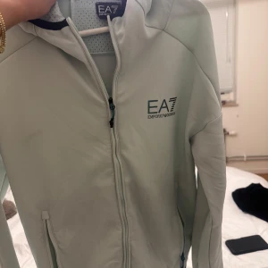 Ljusgrå vindjacka från EA7 Emporio Armani - Snygg ljusgrå vindjacka från EA7 Emporio Armani med huva och dragkedja framtill. Jackan har två fickor med dragkedja och EA7-logga på bröstet. Perfekt för dig som vill ha en stilren och sportig look.