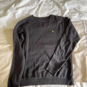 Svart sweatshirt från Lyle & Scott - Svart sweatshirt från Lyle & Scott med klassisk rund hals och gul logga broderad på bröstet. Tröjan har ribbade muddar vid ärmslut och nederkant, samt lång ärm. Perfekt för en stilren och enkel look.