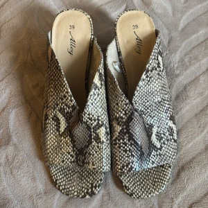Ormskinnsmönstrade sandaler med klack - Säljer ett par sandaler från Alley i storlek 39 med ormskinnsmönster i beige och bruna toner. Skorna har öppen tå, bred klack och en snygg omlott-design framtill. Perfekta för dig som vill sticka ut med ett djurmönster och gillar chunky klackar.