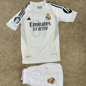 Real Madrid Mbappé fotbollströja set - Säljer ett Real Madrid fotbollströja och shorts med Mbappé och nummer 10 på ryggen. Tröjan är vit med svarta detaljer, gula linjer och klubbmärke på bröstet. Adidas-logga och Champions League-märke på ärmen. Tillverkad i lätt polyester för maximal komfort.