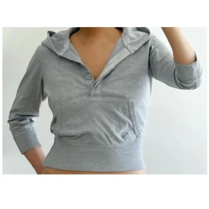 Grå croppad hoodie med huva - Snygg grå croppad hoodie med huva och trekvartsärm. Tröjan har en knapp framtill och en stor ficka på magen. Perfekt för en chill och avslappnad look. Storlek L, passar S/M