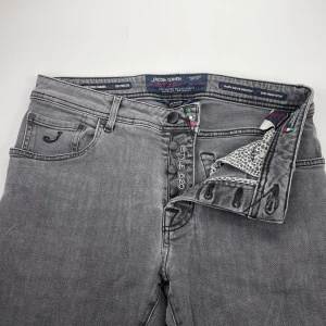 Jacob Cohën Style 620 - Snygga grå jeans från Jacob Cohën, modell Style 620. Klassisk femficksdesign med svarta knappar och unika detaljer på insidan. Jeansen har en diskret broderad logga på fickan och svart patch bak. Perfekta för dig som gillar stilrena och moderna jeans.