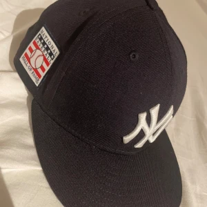 New Era Yankees svart keps, STL 57,7cm (7 1/4) - Svart New Era keps med klassiskt New York Yankees broderat logga i vitt framtill och Hall of Fame-märke på sidan. Snygg böjd skärm och strukturerad passform. Perfekt för dig som gillar sportig streetstyle. 