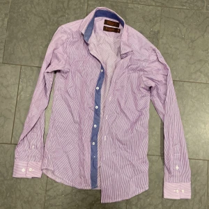 Randig lila skjorta från Bossini - Snygg lila skjorta från Bossini med tunna vita ränder och kontrastfärgad blå insida på kragen. Skjortan har lång ärm, klassisk krage och knäppning framtill. Tillverkad i en mix av bomull och polyester för en skön känsla.