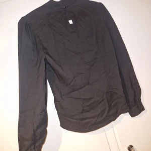 Skjorta / blus - Stilren svart skjorta med lång ärm och hög hals. Skjortan har knappar vid halsen och en rak passform. Materialet känns lätt och har en slät yta. Perfekt för dig som gillar minimalistisk stil och vill ha en enkel men snygg look.