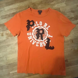 Orange t-shirt Polo Ralph Lauren L - Orange t-shirt från Polo Ralph Lauren med stort vitt tryck och svarta filtapplikationer på bröstet. Klassisk rund hals och korta ärmar. Skön bomullskvalitet och cool universitetsstil. Perfekt för dig som gillar streetwear och sportig vibe.
