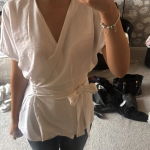 Vit stradivarius omlottblus med knyt i midjan - Säljer en fräsch vit omlottblus med korta ärmar och snygg v-ringning. Blusen har ett brett knytband i midjan som ger en cool siluett och extra detalj. Materialet känns mjukt och har ett lätt fall. Perfekt för dig som gillar stilrena och trendiga plagg.