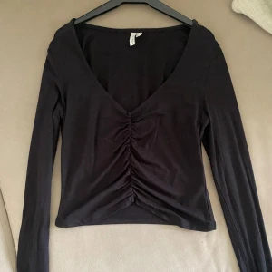 Svart långärmad croppad topp - Svart långärmad topp från NLY Trend med v-ringning och snygg rynkdetalj framtill. Modellen är croppad och har en tight passform som framhäver siluetten. Perfekt att matcha med höga jeans eller kjol.