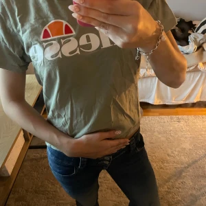 Grön t-shirt från Ellesse - Säljer en grön t-shirt från Ellesse med stor vit och orange logga framtill. Klassisk passform med korta ärmar och rund hals. Tillverkad i mjuk bomull som känns skön mot huden. Perfekt för en avslappnad och sportig stil. Har klippt av nertill och sytt om med en egen söm