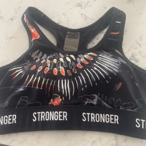 Svart sporttopp från Stronger XS - Sporttopp från Stronger i svart med färgstarkt abstrakt mönster i orange, vitt och grått. Resårband med loggan STRONGER runt om. Racerback med öppning i ryggen. Tillverkad i återvunnen polyester och elastan för en stretchig och bekväm passform. Hör av er vid frågor! Pris kan alltid diskuteras💗