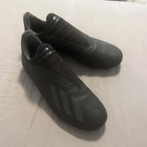 Svarta adidas fotbollsskor  - Säljer ett par svarta adidas fotbollsskor med stilrena, matta ränder på sidan och en modern design utan snörning. Ovandelen är i syntetmaterial med textilinslag och sulan har fasta dobbar för spel på gräs. 