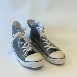 Grå höga sneakers med vit sula - Säljer ett par klassiska höga sneakers i grått med vit gummisula och vit tå. Skorna har snörning med vita skosnören och kontrasterande vita sömmar. Yttermaterialet är bomullstyg och sulan är svart undertill med rutmönster för extra grepp.