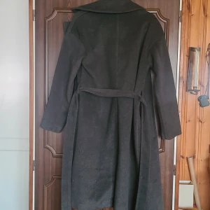 Mörkgrå kappa med bälte från Amisu - Snygg mörkgrå kappa från Amisu med klassisk krage och knytskärp i midjan. Lång modell med slits bak och två fickor framtill. Perfekt för kyligare dagar och ger en clean look till alla outfits. 