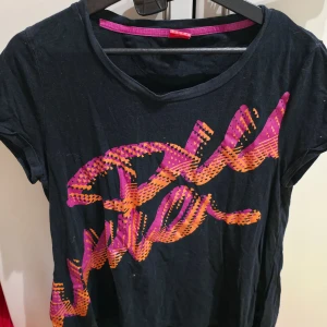 Svart Puma t-shirt med tryck XL - Svart t-shirt från Puma i storlek XL med ett stort grafiskt tryck i orange och rosa över framsidan. Rund halsringning och korta ärmar. Materialet är mjuk bomull, perfekt för en sportig och avslappnad stil. Snygg kontrast med färgstarka detaljer.