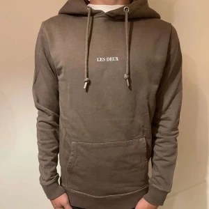 Brun hoodie från Les Deux - Säljer en brun hoodie från Les Deux med vit broderad logga på bröstet. Tröjan har huva med snörning, magficka och ribbade muddar. Perfekt för chill dagar och enkel att matcha med jeans eller joggers.