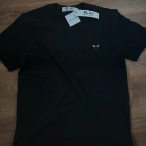 Svart Comme des Garçons Play t-shirt - Svart t-shirt från Comme des Garçons Play med det ikoniska hjärta med ögon broderat på bröstet. Klassisk rund hals och korta ärmar. Tillverkad i 100% bomull för en skön och mjuk känsla. Perfekt för dig som gillar stilrena och trendiga plagg. Du vet varför den är billig